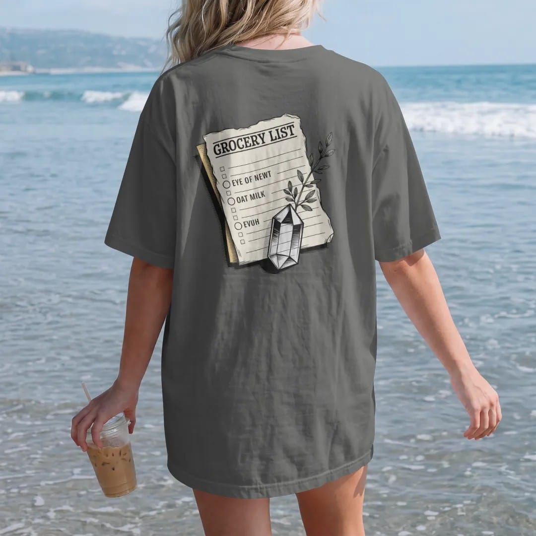 Woman on the beach in a Pepper tee, back view: back print: A torn vintage paper note held down by a small quartz crystal, with a sprig of thyme, list layout with header "GROCERY LIST" and the lines "EYE OF NEWT" and "OAT MILK", detailed vintage…