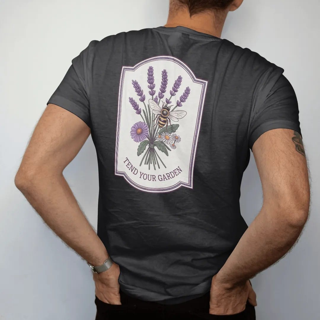 Man wearing a Black tee, seen from the back: back print: A honeybee hovering over a loose bouquet of lavender, calendula and chamomile, small "TEND YOUR GARDEN" beneath the bouquet, antique apothecary-label engraving, fine crosshatching, ornate…