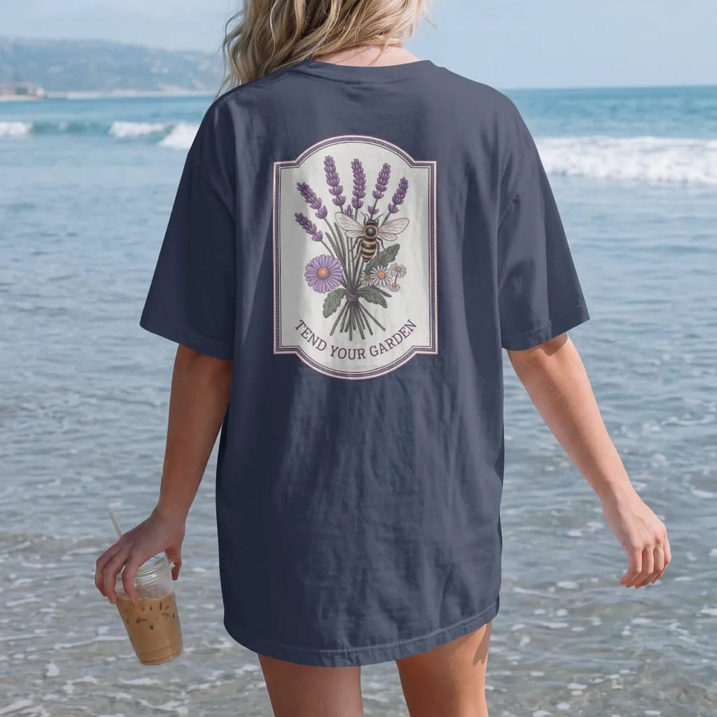 Woman on the beach in a Navy tee, back view: back print: A honeybee hovering over a loose bouquet of lavender, calendula and chamomile, small "TEND YOUR GARDEN" beneath the bouquet, antique apothecary-label engraving, fine crosshatching, ornate…