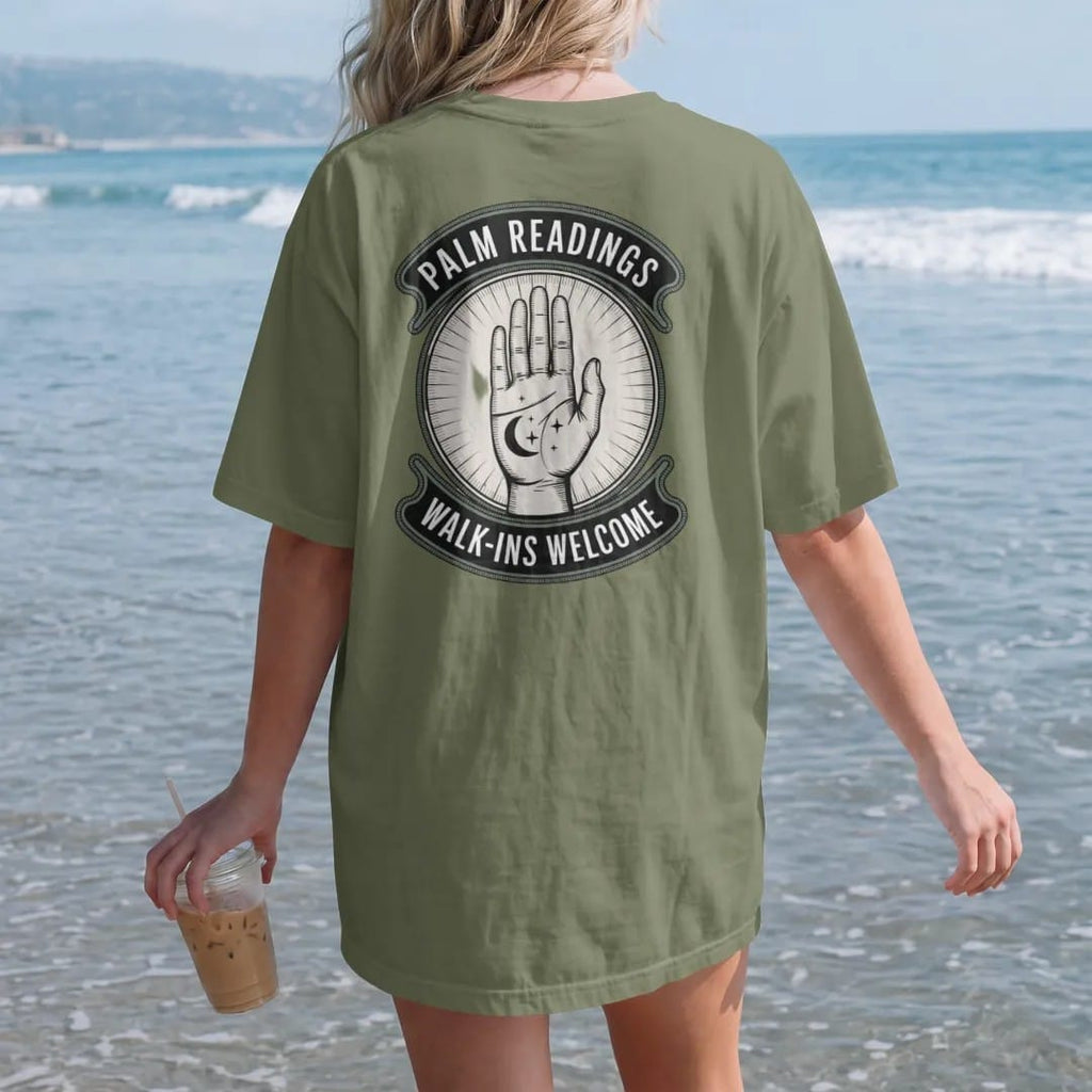 Woman on the beach in a Moss tee, back view: back print: an open palm with fine lines drawn in and a crescent moon and stars at the fingertips, vintage storefront sign with "PALM READINGS" on top and "WALK-INS WELCOME" below, detailed vintage…