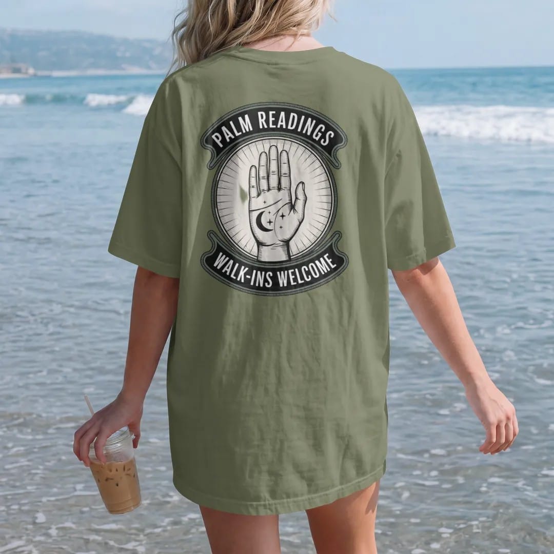 Woman on the beach in a Moss tee, back view: back print: an open palm with fine lines drawn in and a crescent moon and stars at the fingertips, vintage storefront sign with "PALM READINGS" on top and "WALK-INS WELCOME" below, detailed vintage…