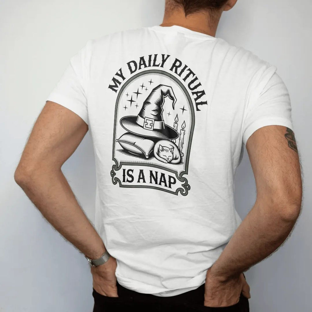 Man wearing a White tee, seen from the back: back print: A witch hat resting on a pillow with a sleeping cat curled against it and candles nearby, arched "MY DAILY RITUAL" above and "IS A NAP" below, antique apothecary-label engraving, fine…