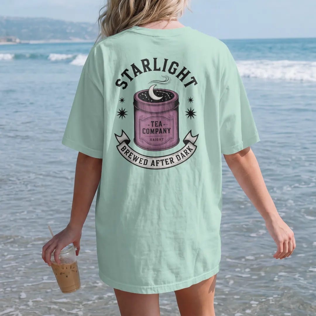 Woman on the beach in a Chalky Mint tee, back view: back print: A vintage tea tin with a crescent moon and constellations on the lid and steam curling up, "STARLIGHT" arched above, "TEA COMPANY" on the tin and "BREWED AFTER DARK" in a ribbon below,…