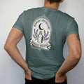 Man wearing a Blue Spruce tee, seen from the back: back print: an ornate crest with a crescent moon, a corked bottle and crossed sprigs of lavender and rosemary, crest with "MIDNIGHT APOTHECARY" arched above and "REMEDIES & RITUALS" in a ribbon…