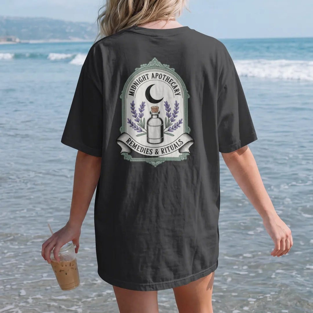 Woman on the beach in a Black tee, back view: back print: an ornate crest with a crescent moon, a corked bottle and crossed sprigs of lavender and rosemary, crest with "MIDNIGHT APOTHECARY" arched above and "REMEDIES & RITUALS" in a ribbon below,…