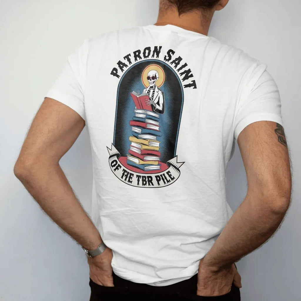 Man wearing a White tee, seen from the back: a haloed skeleton librarian in cat-eye glasses on a tall stack of books in a prayer-candle frame, under Patron Saint and over a banner reading Of The TBR Pile, in pulp halftone cream, red, yellow and blue.
