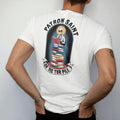 Man wearing a White tee, seen from the back: a haloed skeleton librarian in cat-eye glasses on a tall stack of books in a prayer-candle frame, under Patron Saint and over a banner reading Of The TBR Pile, in pulp halftone cream, red, yellow and blue.