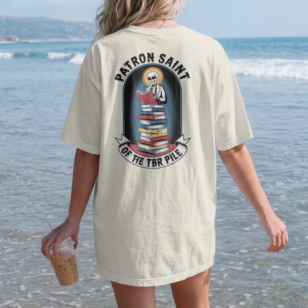 Woman on the beach in a Ivory tee, back view: a haloed skeleton librarian in cat-eye glasses on a tall stack of books in a prayer-candle frame, under Patron Saint and over a banner reading Of The TBR Pile, in pulp halftone cream, red, yellow and…