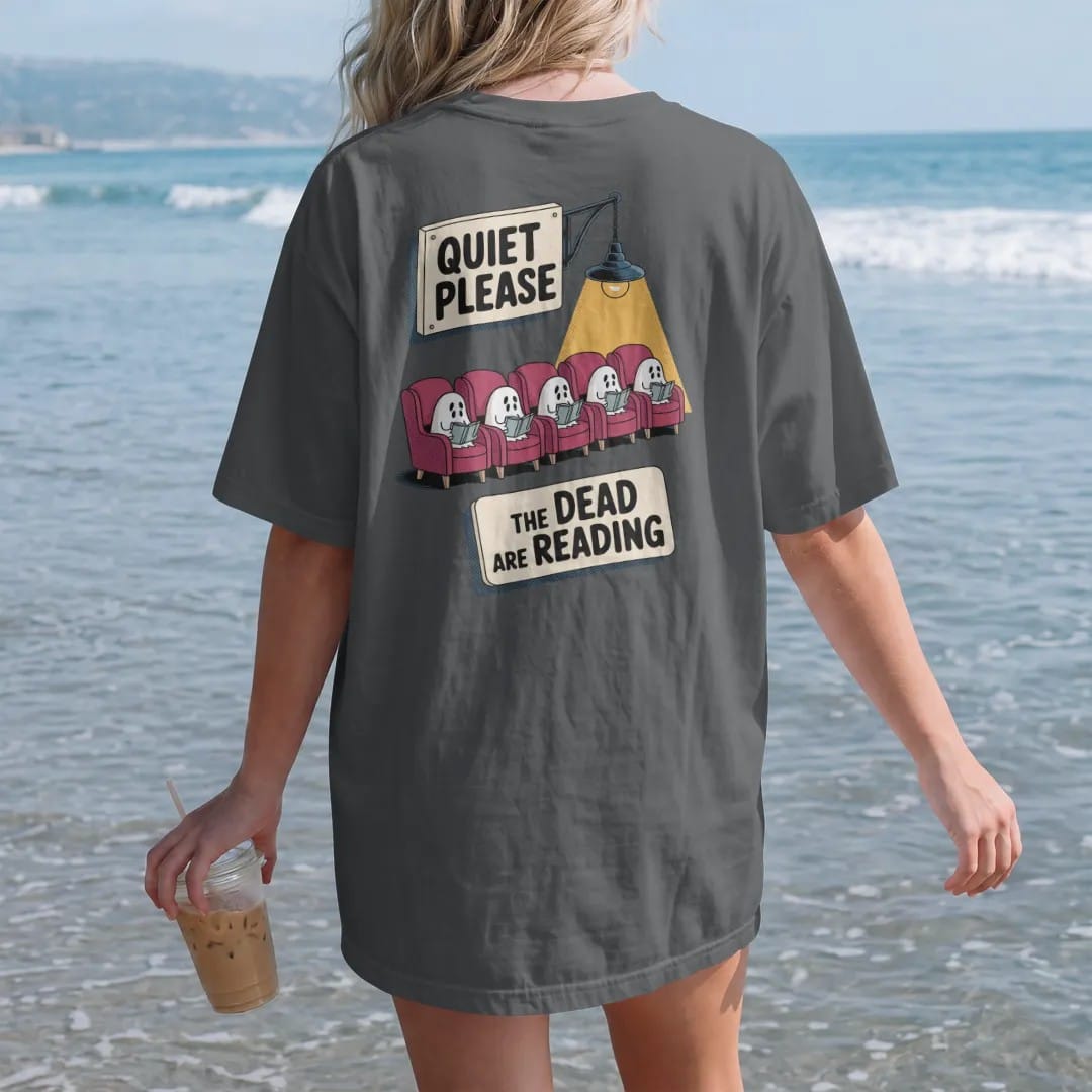Woman on the beach in a Graphite tee, back view: a library placard reading Quiet Please over a row of small ghosts reading in armchairs, with The Dead Are Reading below, in pulp halftone cream, red, yellow and blue.