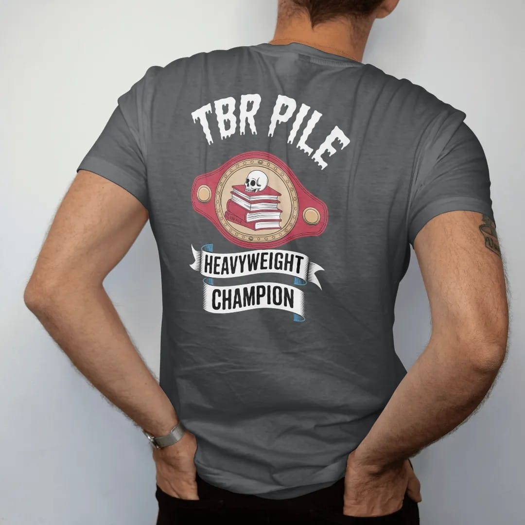 Man wearing a Graphite tee, seen from the back: a prize-fight championship belt with a book stack and skull on the center plate reading TBR Pile and a Heavyweight Champion ribbon, in pulp halftone cream, red, yellow and blue.