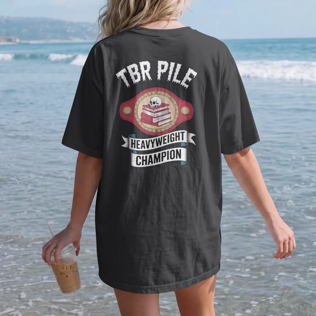 Woman on the beach in a Black tee, back view: a prize-fight championship belt with a book stack and skull on the center plate reading TBR Pile and a Heavyweight Champion ribbon, in pulp halftone cream, red, yellow and blue.