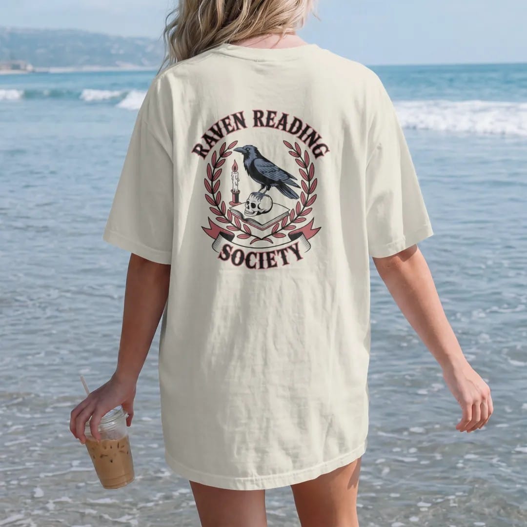 Woman on the beach in a Ivory tee, back view: a crest with a raven perched on a book and skull, Raven Reading arched above and a Society ribbon below, in pulp halftone cream, red, yellow and blue.