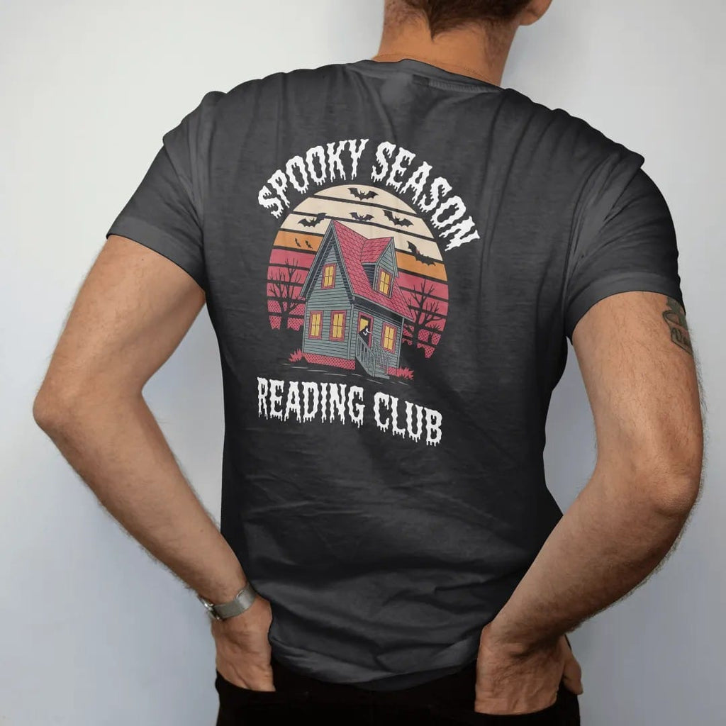 Man wearing a Black tee, seen from the back: a haunted house with one lit attic window over retro sunset stripes with bats and leaves, Spooky Season arched above and Reading Club below, in pulp halftone cream, red, yellow and blue.