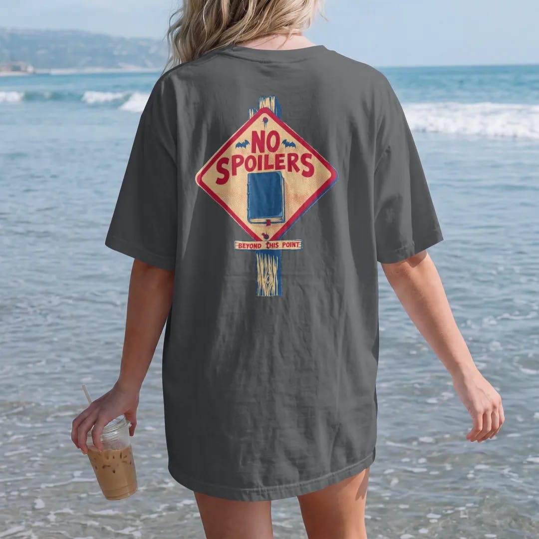 Woman on the beach in a Graphite tee, back view: a diamond caution sign with a tombstone and closed book reading No Spoilers on a graveyard post, with Beyond This Point on a plank, in pulp halftone cream, red, yellow and blue.