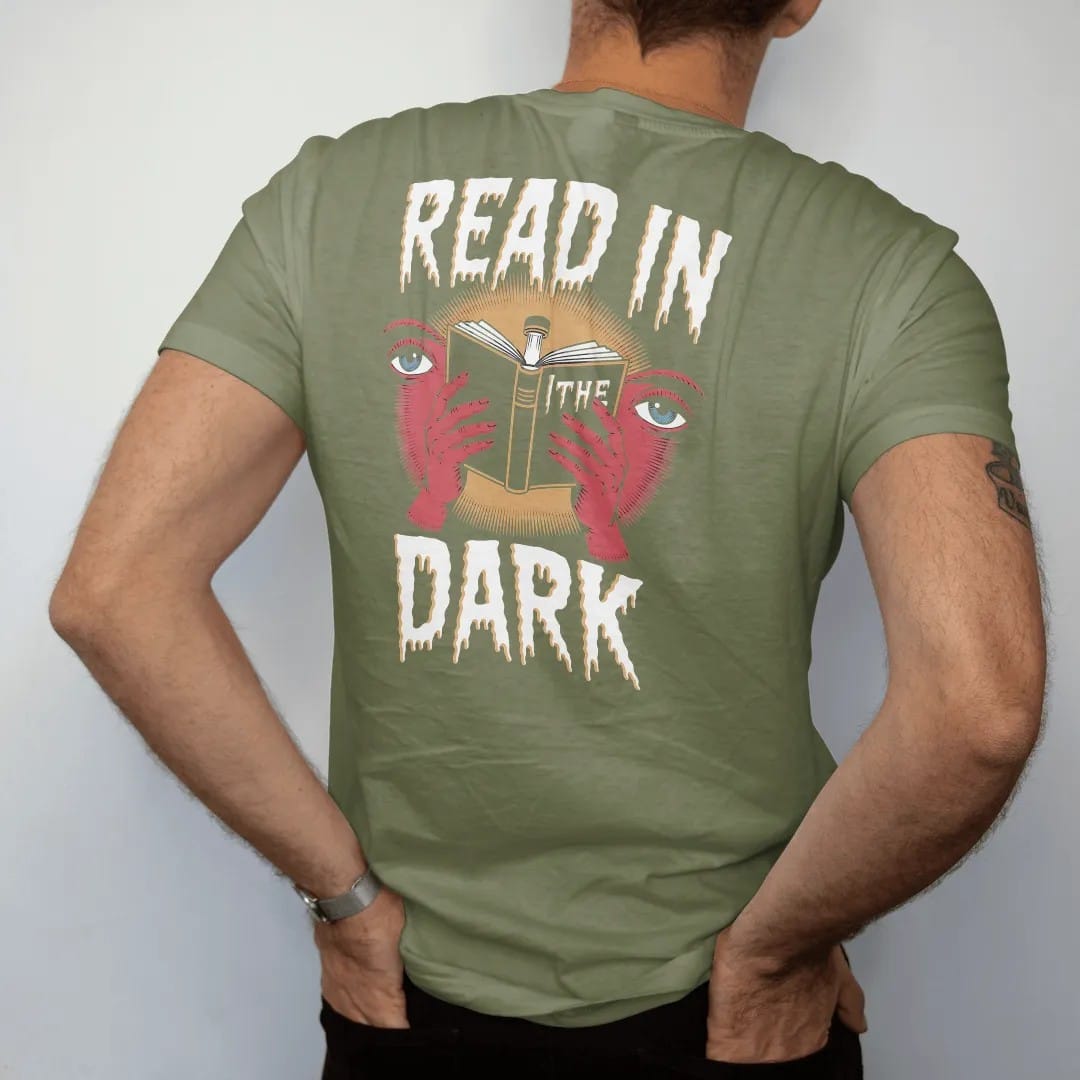 Man wearing a Moss tee, seen from the back: an open book glowing from a clip-on book light with eyes watching in the shadows, with Read In above and The Dark below, in pulp halftone cream, red, yellow and blue.