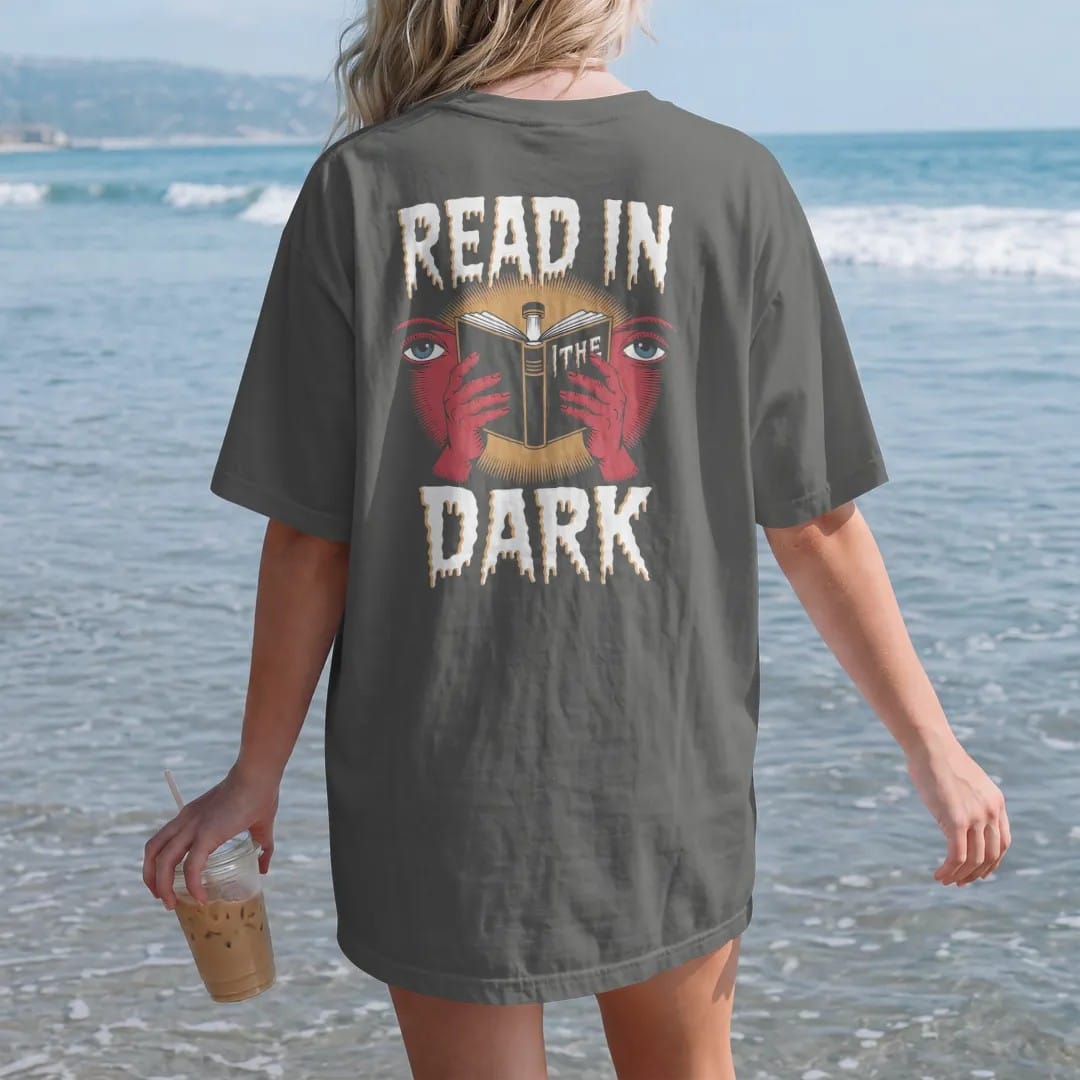 Woman on the beach in a Pepper tee, back view: an open book glowing from a clip-on book light with eyes watching in the shadows, with Read In above and The Dark below, in pulp halftone cream, red, yellow and blue.