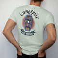 Man wearing a Bay tee, seen from the back: a coffin-shaped bookshelf full of books, candles and a skull with a raven on top, Coffin Shelf arched above and Est. Midnight below, in pulp halftone navy, red, mustard and teal.