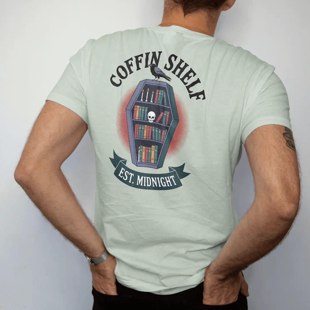 Man wearing a Bay tee, seen from the back: a coffin-shaped bookshelf full of books, candles and a skull with a raven on top, Coffin Shelf arched above and Est. Midnight below, in pulp halftone navy, red, mustard and teal.
