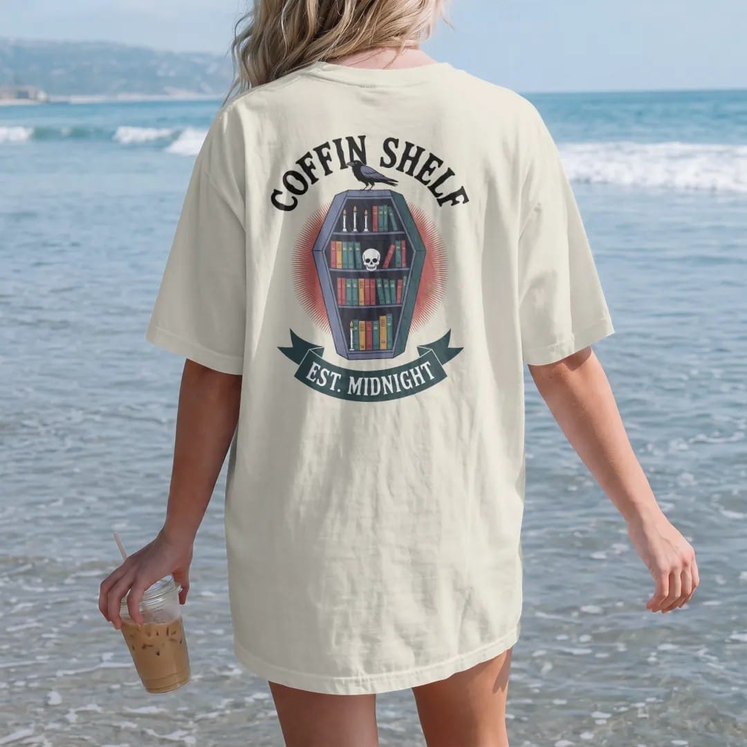 Woman on the beach in a Ivory tee, back view: a coffin-shaped bookshelf full of books, candles and a skull with a raven on top, Coffin Shelf arched above and Est. Midnight below, in pulp halftone navy, red, mustard and teal.