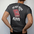 Man wearing a Black tee, seen from the back: a chained grimoire with a staring eye on the cover and smoke curling from the pages, with Do Not Read above and Aloud below, in pulp halftone cream, red, yellow and blue.