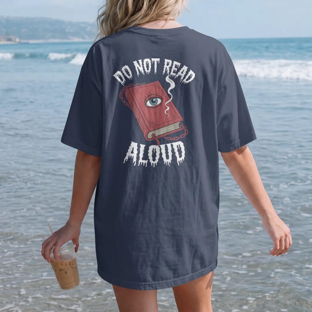 Woman on the beach in a Navy tee, back view: a chained grimoire with a staring eye on the cover and smoke curling from the pages, with Do Not Read above and Aloud below, in pulp halftone cream, red, yellow and blue.