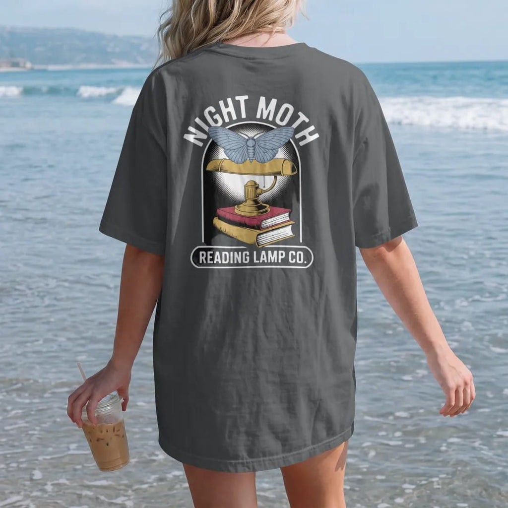 Woman on the beach in a Graphite tee, back view: a large moth circling a brass reading lamp over a book stack, Night Moth arched above and Reading Lamp Co. on a bar below, in pulp halftone cream, red, yellow and blue.