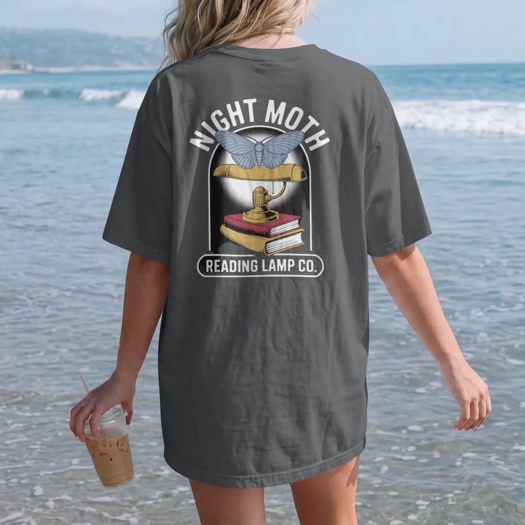 Woman on the beach in a Graphite tee, back view: a large moth circling a brass reading lamp over a book stack, Night Moth arched above and Reading Lamp Co. on a bar below, in pulp halftone cream, red, yellow and blue.