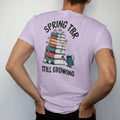 Man wearing a Orchid tee, seen from the back: a tall book stack sprouting vines, mushrooms and skull flowers beside a tipped watering can, Spring TBR above and Still Growing below, in pulp halftone navy, red, mustard and teal.