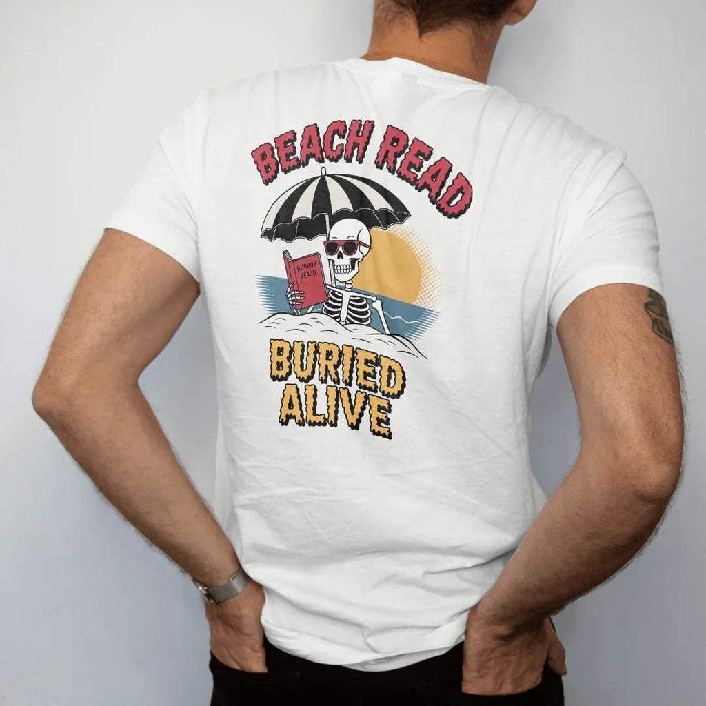 Man wearing a White tee, seen from the back: a skeleton in sunglasses half buried in sand under a beach umbrella reading a horror paperback, with Beach Read above and Buried Alive below, in pulp halftone cream, red, yellow and blue.
