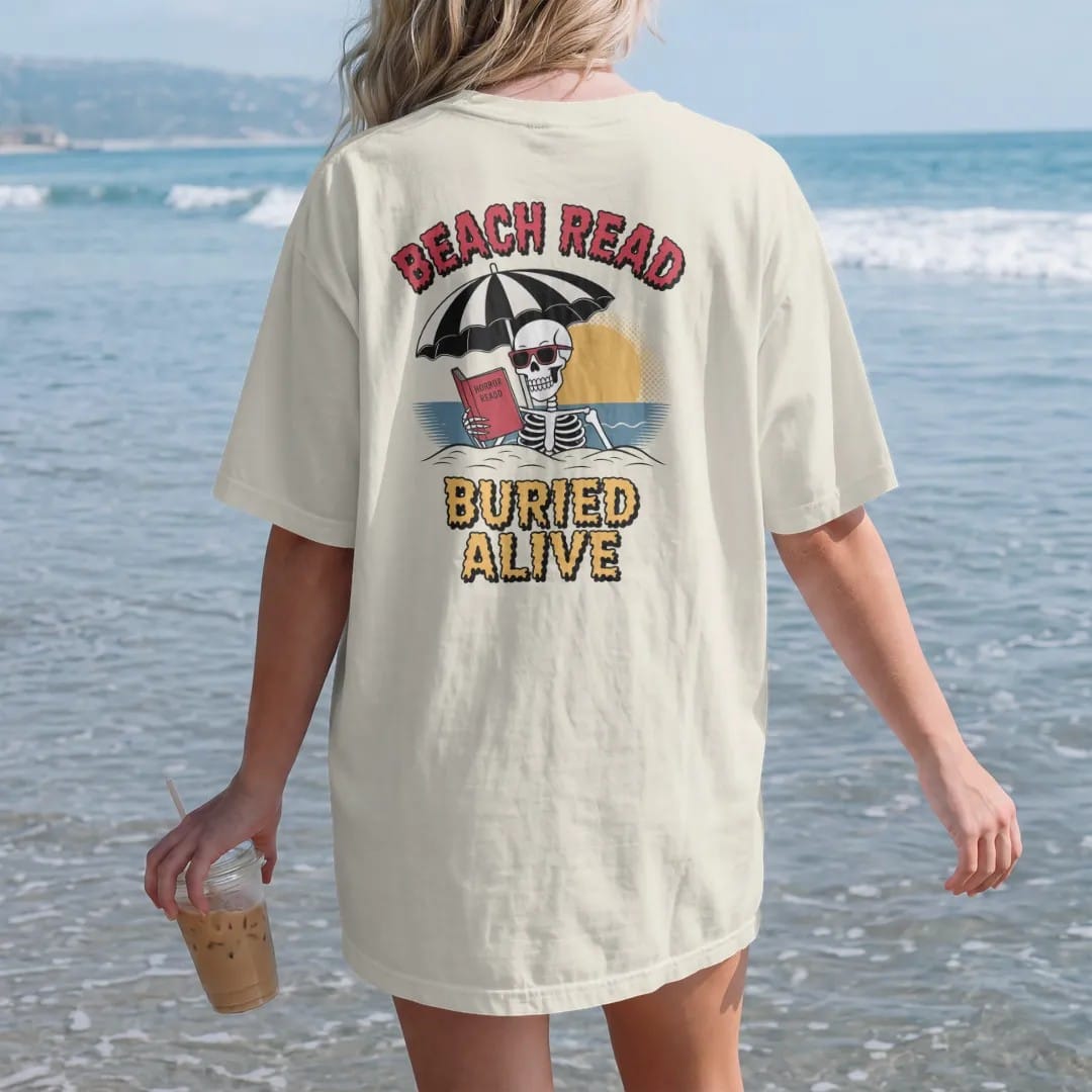 Woman on the beach in a Ivory tee, back view: a skeleton in sunglasses half buried in sand under a beach umbrella reading a horror paperback, with Beach Read above and Buried Alive below, in pulp halftone cream, red, yellow and blue.