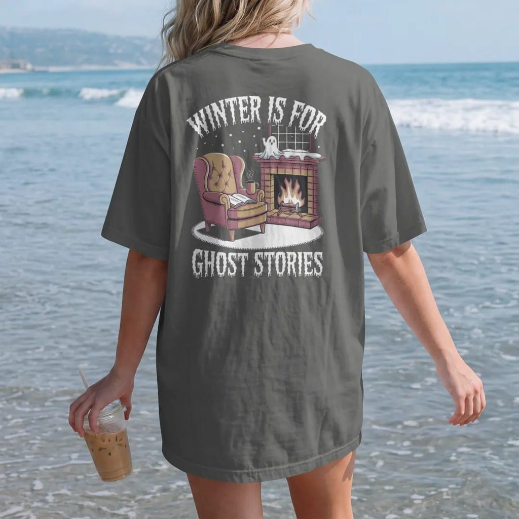 Woman on the beach in a Pepper tee, back view: a wingback armchair by a fireplace with a book, steaming mug and a small ghost warming its hands, with Winter Is For above and Ghost Stories below, in pulp halftone cream, red, yellow and blue.