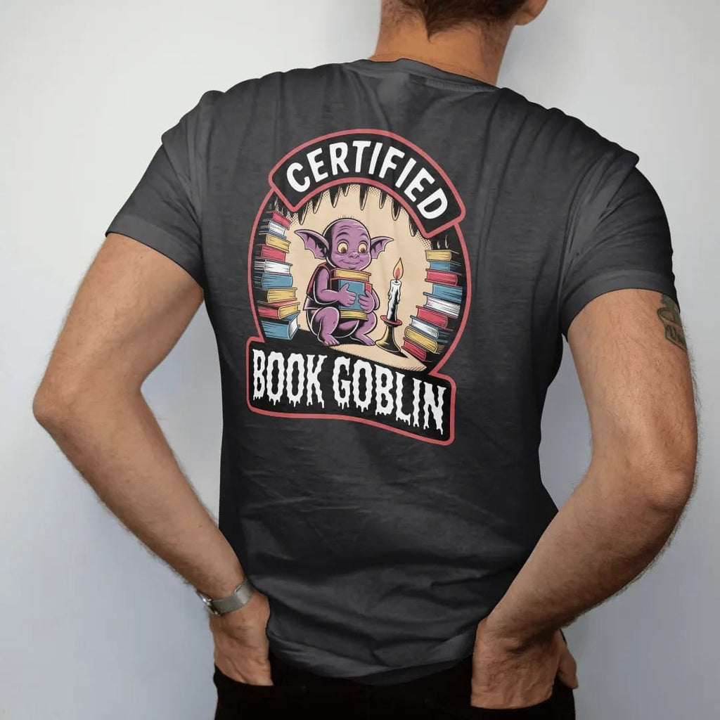Man wearing a Black tee, seen from the back: a goblin hugging an armful of books in a candlelit cave of book piles inside a seal badge reading Certified Book Goblin, in pulp halftone cream, red, yellow and blue.