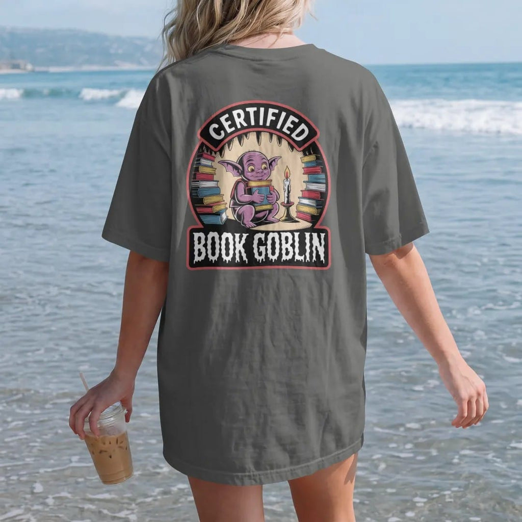 Woman on the beach in a Pepper tee, back view: a goblin hugging an armful of books in a candlelit cave of book piles inside a seal badge reading Certified Book Goblin, in pulp halftone cream, red, yellow and blue.