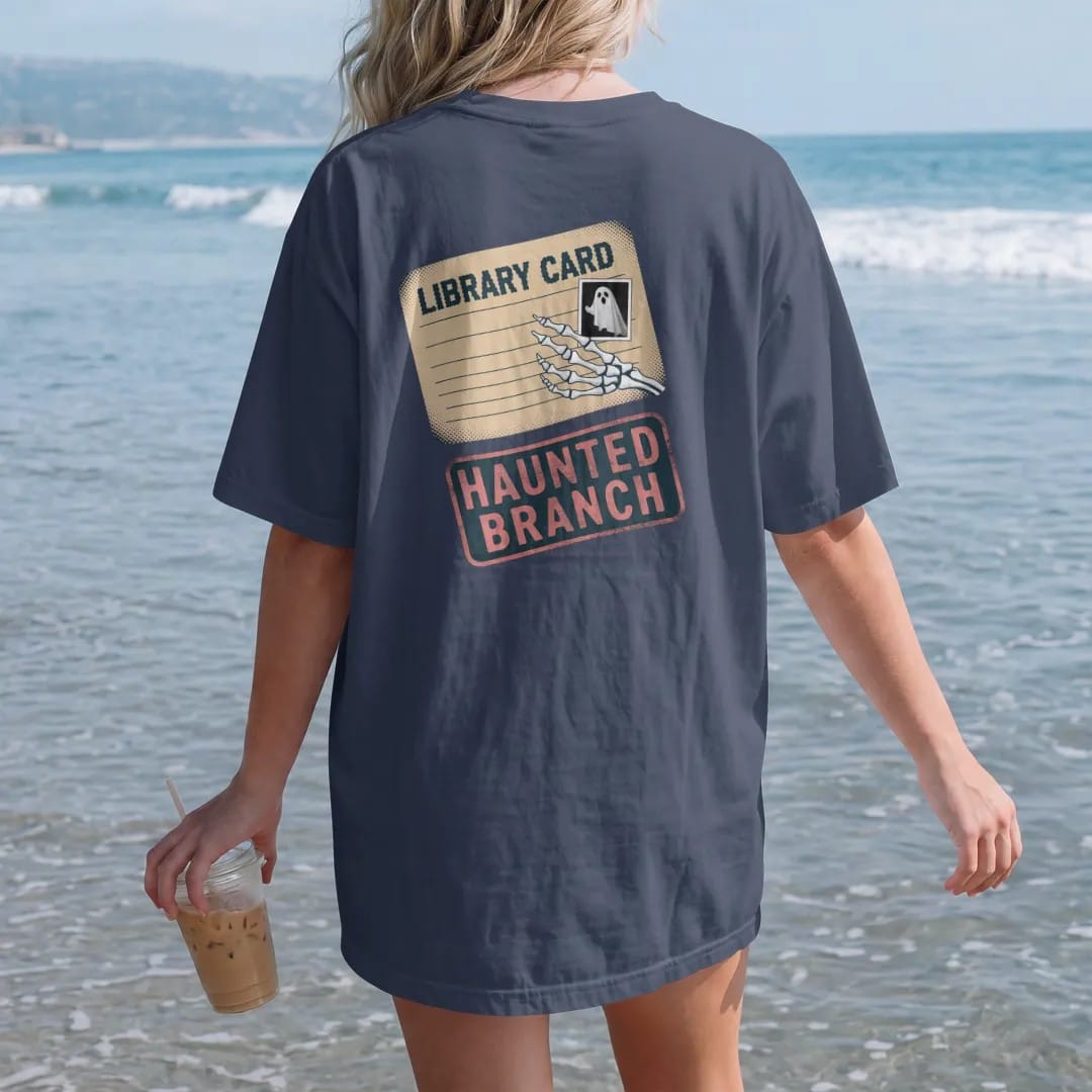 Woman on the beach in a Navy tee, back view: a skeleton hand holding an old library card with a small ghost photo, Library Card across the top and Haunted Branch stamped below, in pulp halftone navy, red, mustard and teal.