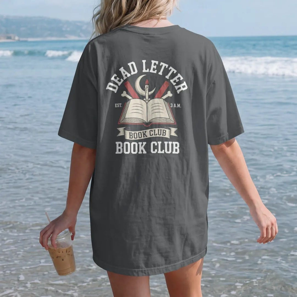 Woman on the beach in a Graphite tee, back view: a round crest with crescent moon, candle and bone bookmarks around an open book, Dead Letter arched above, Book Club below and an Est. 3 A.M. ribbon, in pulp halftone cream, red, yellow and blue.
