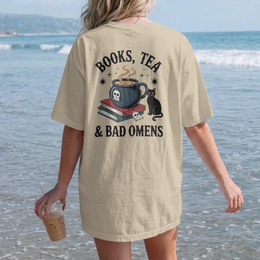 Woman on the beach in a Sandstone tee, back view: a cauldron teacup with a skull tea tag beside a book stack and black cat, Books, Tea arched above and & Bad Omens below, in pulp halftone navy, red, mustard and teal.