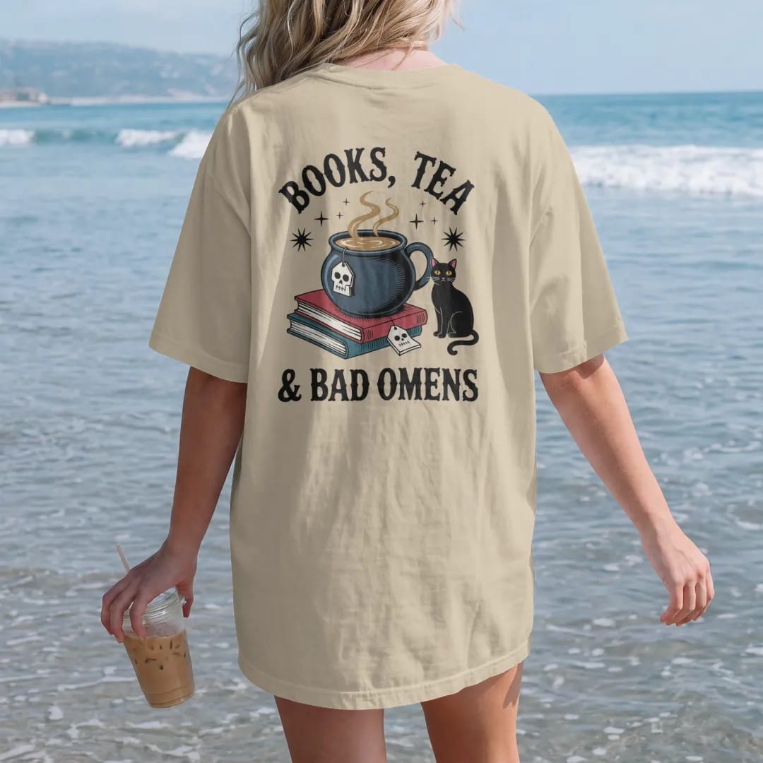 Woman on the beach in a Sandstone tee, back view: a cauldron teacup with a skull tea tag beside a book stack and black cat, Books, Tea arched above and & Bad Omens below, in pulp halftone navy, red, mustard and teal.