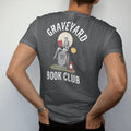 Man wearing a Graphite tee, seen from the back: a skeleton reading on a tombstone by lantern light under a full moon, Graveyard arched above and Book Club below, in pulp halftone cream, red, yellow and blue.