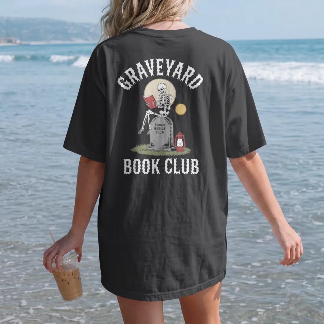 Woman on the beach in a Black tee, back view: a skeleton reading on a tombstone by lantern light under a full moon, Graveyard arched above and Book Club below, in pulp halftone cream, red, yellow and blue.