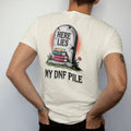 Man wearing a Ivory tee, seen from the back: a cracked tombstone carved Here Lies over a small pile of buried books with My DNF Pile below, in pulp halftone cream, red, yellow and blue.
