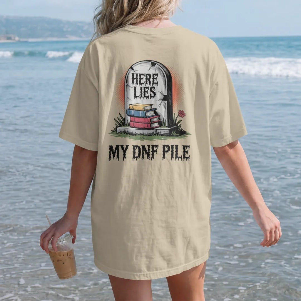 Woman on the beach in a Sandstone tee, back view: a cracked tombstone carved Here Lies over a small pile of buried books with My DNF Pile below, in pulp halftone cream, red, yellow and blue.