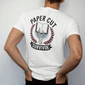Man wearing a White tee, seen from the back: a bandaged skeleton hand holding up an open book inside a laurel wreath, Paper Cut arched above and a Survivor banner below, in pulp halftone cream, red, yellow and blue.