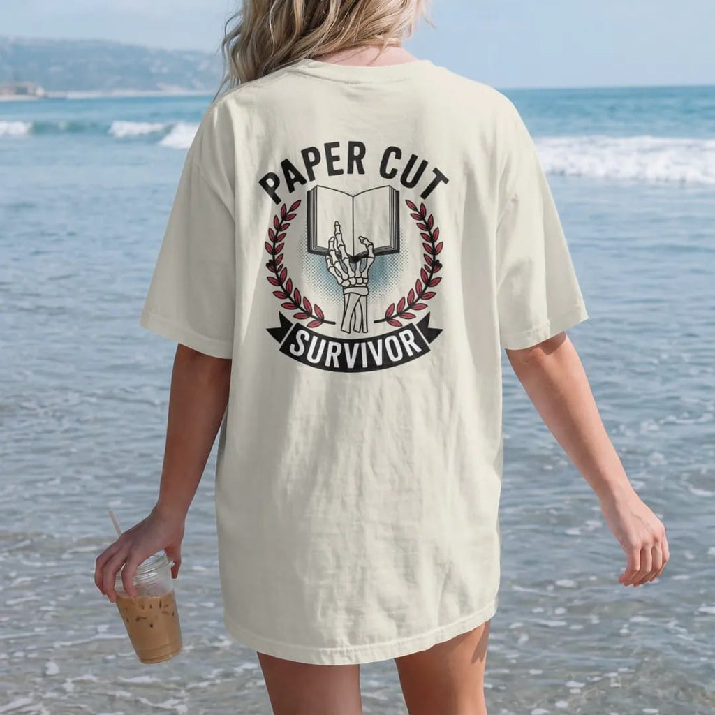 Woman on the beach in a Ivory tee, back view: a bandaged skeleton hand holding up an open book inside a laurel wreath, Paper Cut arched above and a Survivor banner below, in pulp halftone cream, red, yellow and blue.