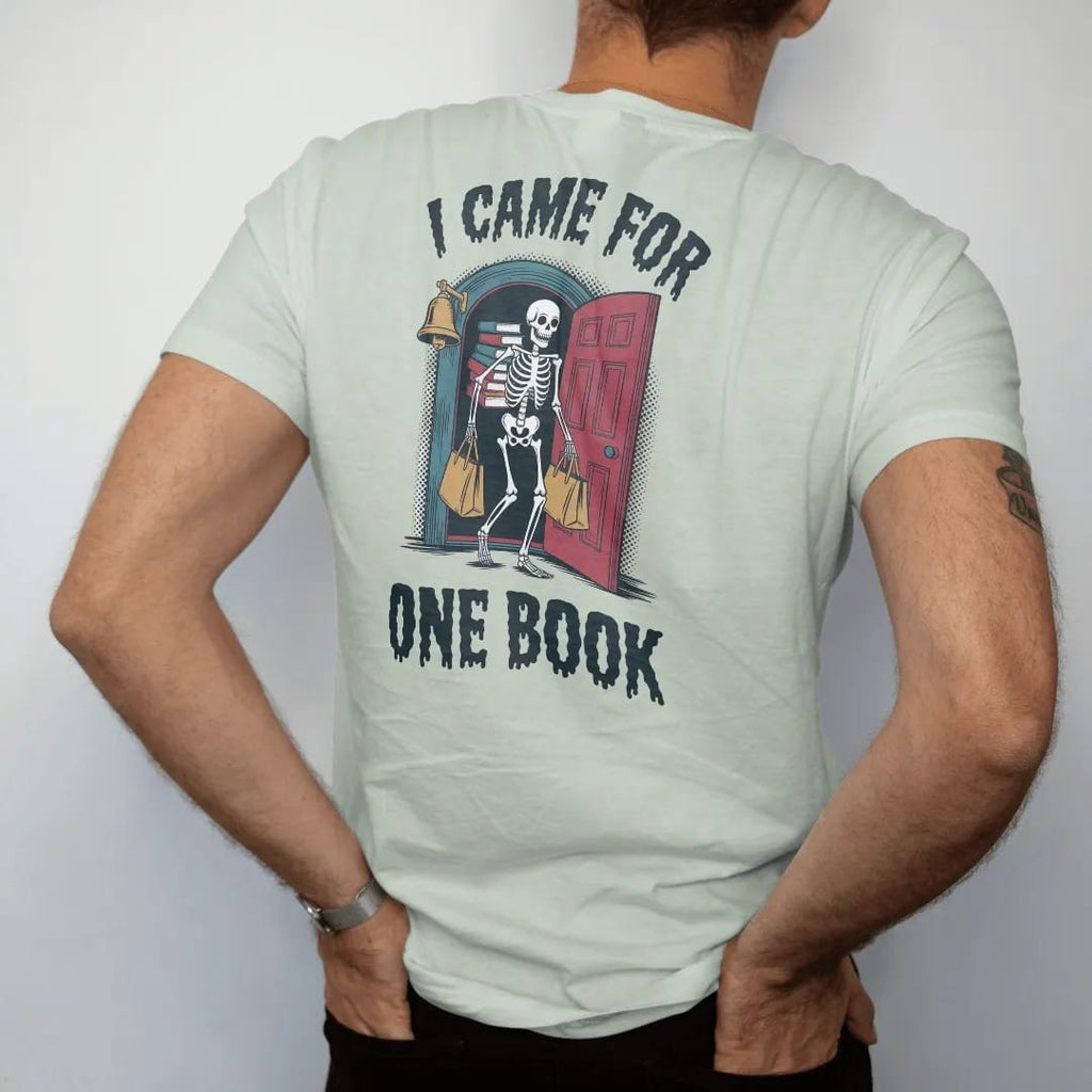 Man wearing a Bay tee, seen from the back: a skeleton leaving an old bookstore carrying a teetering stack of books and two tote bags, with I Came For above and One Book below, in pulp halftone navy, red, mustard and teal.
