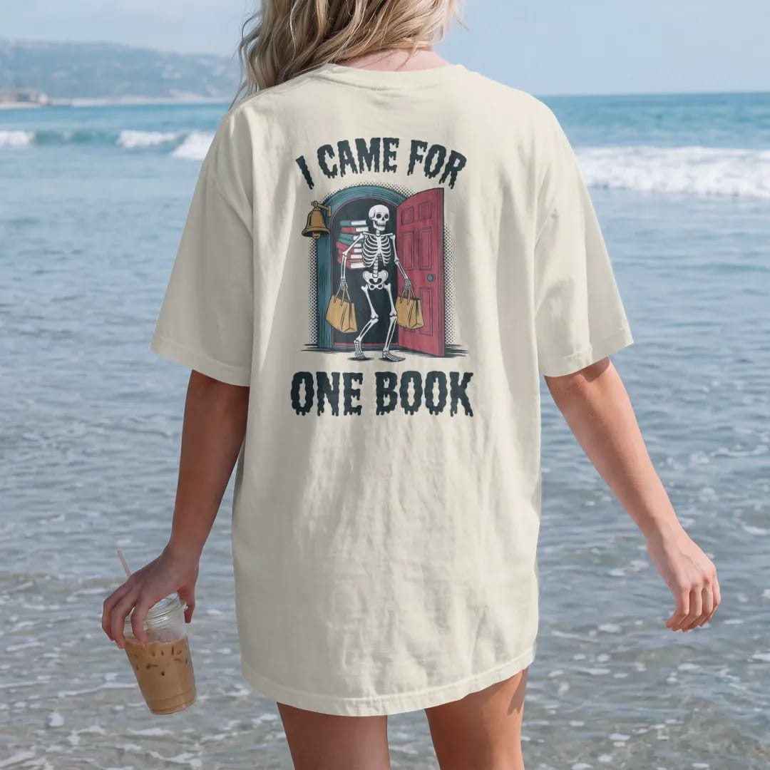 Woman on the beach in a Ivory tee, back view: a skeleton leaving an old bookstore carrying a teetering stack of books and two tote bags, with I Came For above and One Book below, in pulp halftone navy, red, mustard and teal.