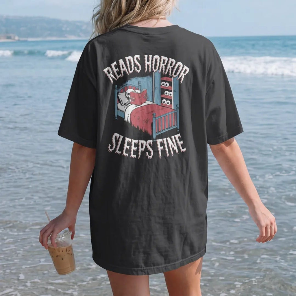 Woman on the beach in a Black tee, back view: a skeleton asleep with a horror book over its face while eyes peek from under the bed and closet, with Reads Horror above and Sleeps Fine below, in pulp halftone cream, red, yellow and blue.