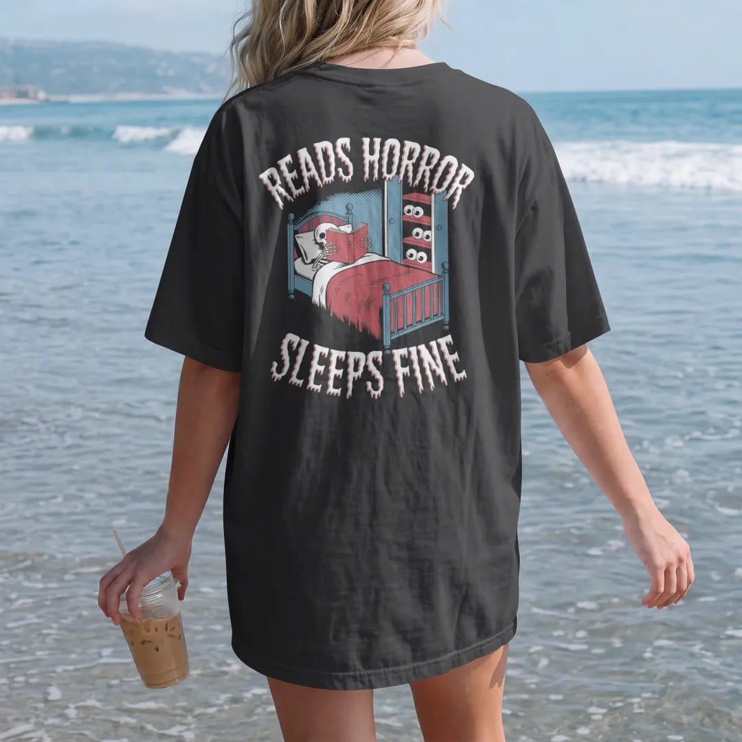 Woman on the beach in a Black tee, back view: a skeleton asleep with a horror book over its face while eyes peek from under the bed and closet, with Reads Horror above and Sleeps Fine below, in pulp halftone cream, red, yellow and blue.