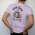 Man wearing a Orchid tee, seen from the back: a vintage apothecary ad with a skull-labeled tonic bottle and books, Book Tonic above and a Cures Daylight ribbon below, in pulp halftone navy, red, mustard and teal.