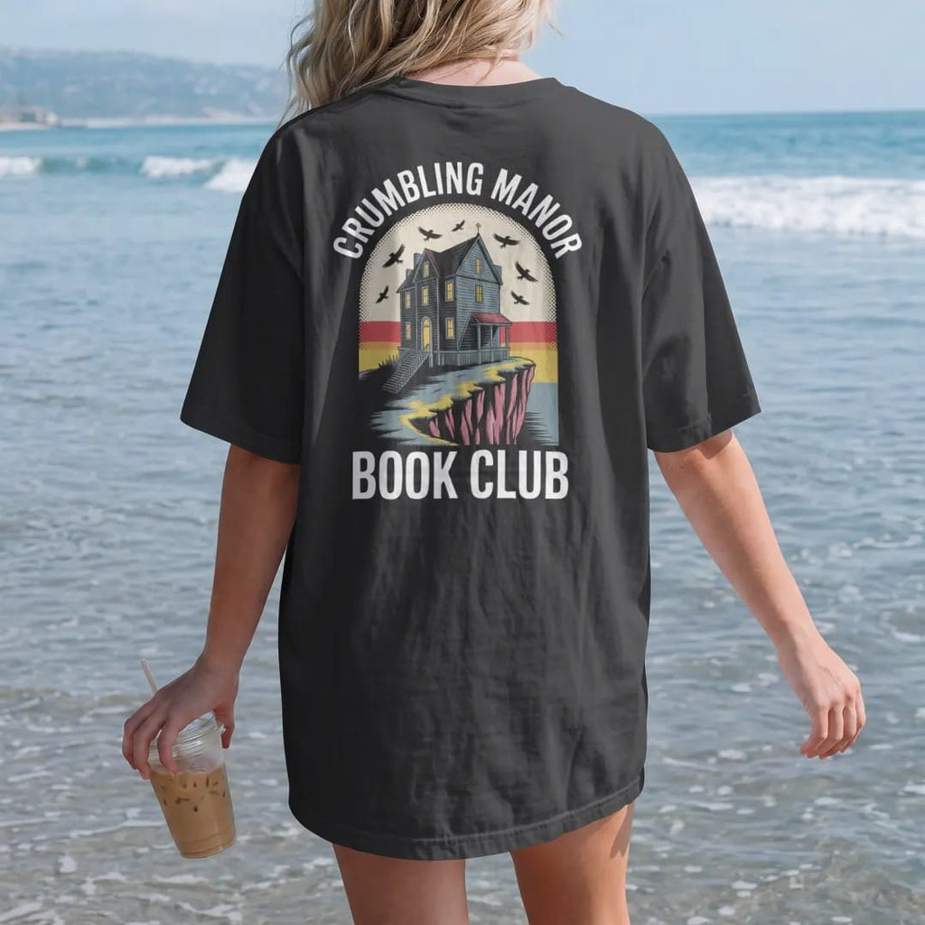 Woman on the beach in a Black tee, back view: a crumbling gothic manor on a cliff with one lit window and ravens circling, Crumbling Manor arched above and Book Club below, in pulp halftone cream, red, yellow and blue.