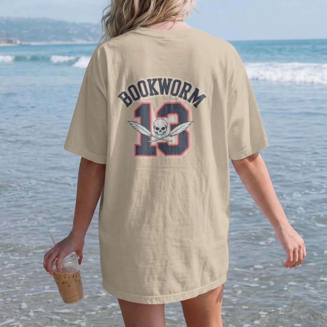 Woman on the beach in a Sandstone tee, back view: a jersey-style Bookworm arch over a large number 13 with a small skull and crossed quills, in pulp halftone cream, red, yellow and blue.